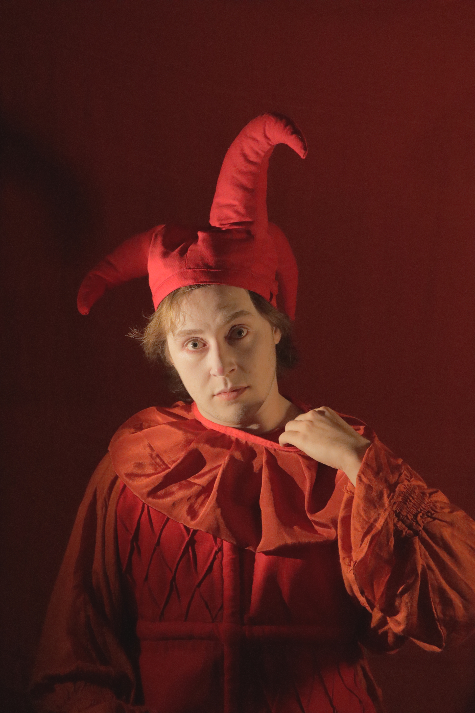 Yorick pulling on the collar of their red jester outfit, staring ahead with a piercing look. 